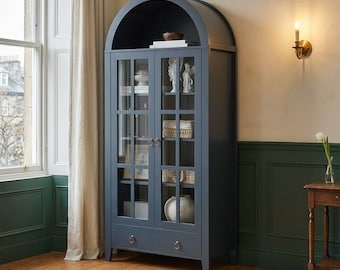 May include: A tall, dark blue cabinet with an arched top and two glass-paned doors. The cabinet features a single drawer at the bottom with two circular metal pulls. Inside, multiple shelves hold various decorative items, including white figurines.