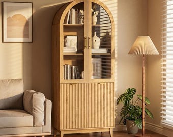 May include: A light wood-toned arched cabinet with glass doors and ribbed lower doors. The cabinet is filled with books, decorative vases, and framed photos. A floor lamp with a pleated shade and a potted plant are nearby.