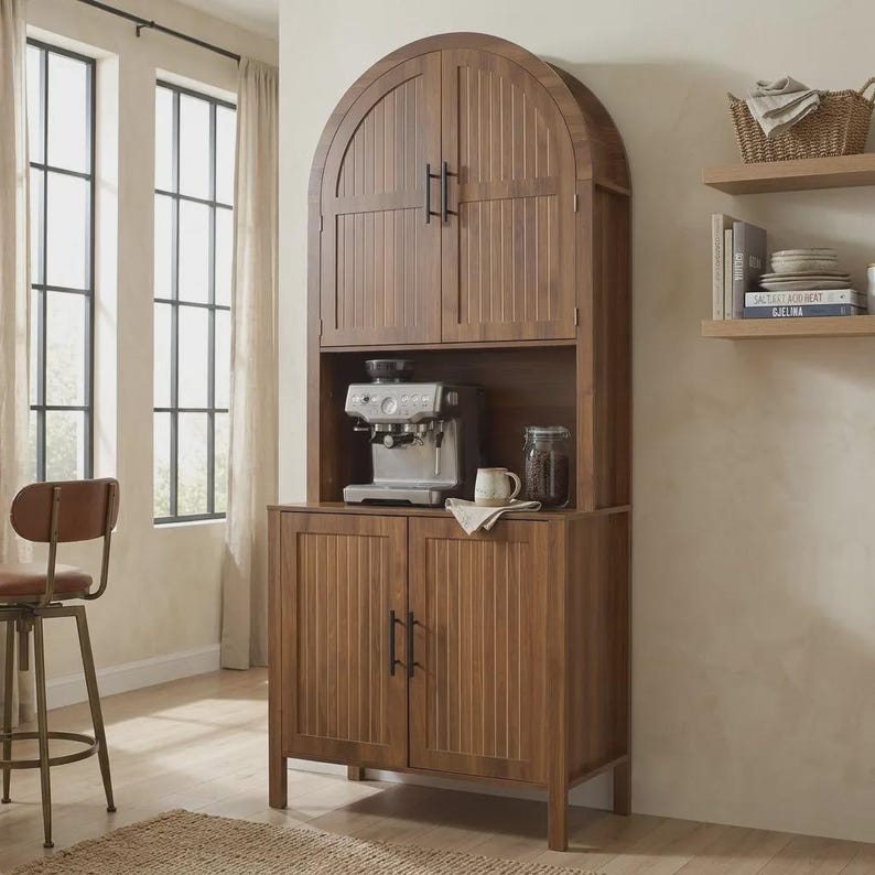 May include: A tall, brown wooden hutch cabinet featuring an arched top, two upper doors with vertical slats, and two lower doors with matching slats. The open middle shelf holds a silver espresso machine, a ceramic mug, and a glass jar.