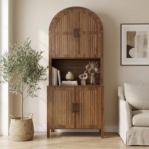 May include: A tall, arched wooden cabinet featuring two upper doors and two lower doors, all with vertical slat detailing and black metal handles. The open middle shelf displays several books, a small ceramic vase, and a decorative sculpture.