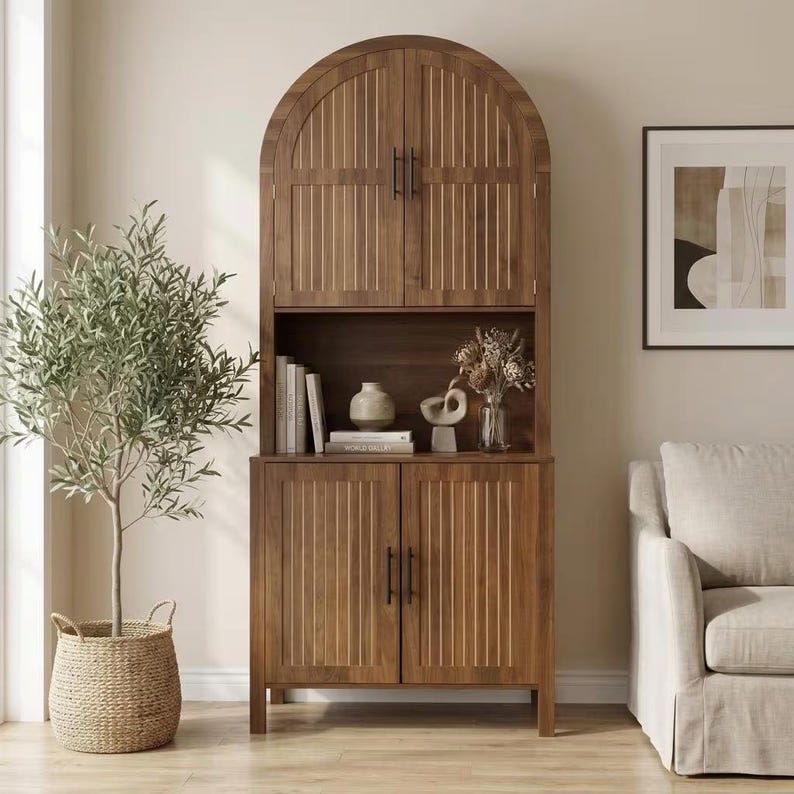 May include: A tall, arched wooden cabinet featuring two upper doors and two lower doors, all with vertical slat detailing and black metal handles. The open middle shelf displays several books, a small ceramic vase, and a decorative sculpture.