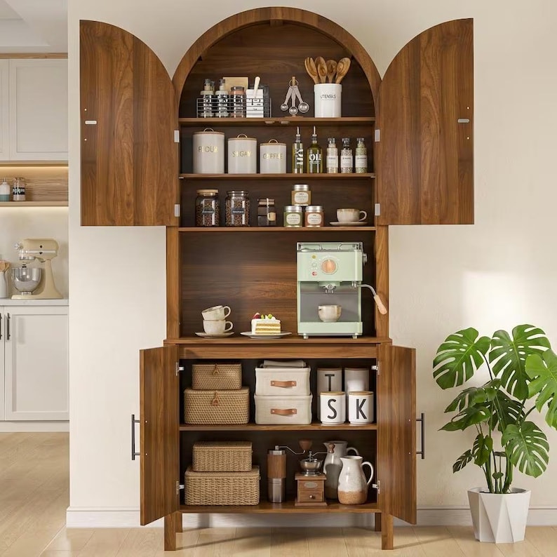 May include: A tall, arched wooden cabinet with open doors, featuring multiple shelves organized with kitchen canisters, a light green espresso machine, woven storage baskets, and various ceramic mugs, jars, and wooden kitchen utensils.