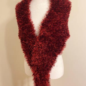 Puede incluir: Bufanda borgoña, esponjosa, exhibida en un maniquí. La bufanda tiene una apariencia suave y texturizada con fibras largas y difusas. La bufanda está drapeada para mostrar su forma y diseño, adecuada para el clima frío.