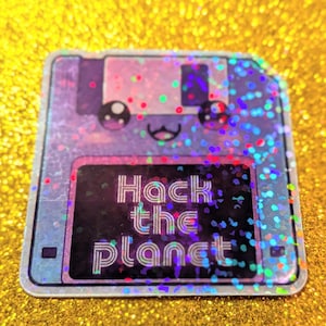 Op de afbeelding: Een holografische sticker in de vorm van een diskette, met een kawaii-gezicht en de tekst "Hack the Planet" in het wit. De sticker heeft een zilveren rand en is geplaatst tegen een gouden glitterachtergrond.