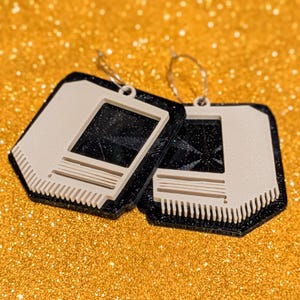 May include: A pair of retro-style earrings. Each earring features a black, square-shaped frame with a cream-colored interior and a rectangular opening. The earrings have a textured, glittery appearance and are suspended from silver-colored hoops.