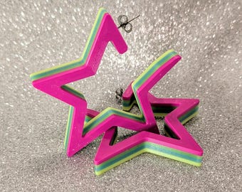 May include: A pair of star-shaped earrings in a vibrant color scheme. The earrings feature a layered design with a hot pink outline, a light green middle layer, and a blue inner layer. The earrings are on a silver glitter background.