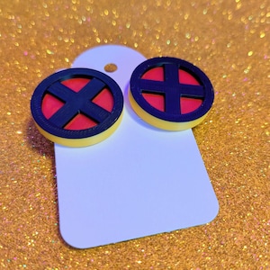 Puede incluir: Un par de pendientes redondos con el logotipo de X-Men. Los pendientes tienen un diseño de X negro sobre un fondo rojo, con un borde amarillo. Se muestran en una etiqueta blanca sobre un fondo dorado brillante.