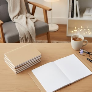 Könnte beinhalten: Ein Stapel beiger Notizbücher mit weiß linierten Seiten liegt neben einem offenen Notizbuch auf einem Holztisch. Eine Tasse Kaffee dampft in der Nähe, mit Brille und Stift. Ein gemütlicher Stuhl und ein Bücherregal sind im Hintergrund.