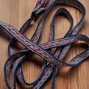 Peut inclure: Ruban tissé avec un motif répétitif de losanges rouges et blancs sur fond bleu. Le ruban est bouclé et noué, avec des extrémités effilochées. Il est posé sur une surface en bois.