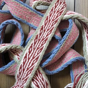 Puede incluir: Primer plano de cinta tejida en tonos rosa, rojo, azul y blanco. La cinta tiene un patrón intrincado con una apariencia texturizada. La cinta está en bucle y entrelazada, creando una exhibición visualmente atractiva. La cinta probablemente se utiliza para manualidades o fines decorativos.