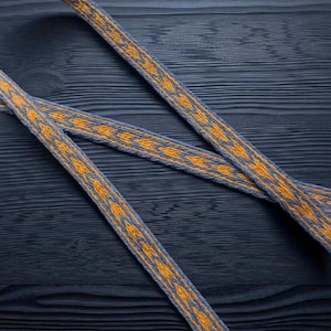 May include: Close-up of three woven ribbons with a repeating geometric pattern. The ribbons are blue with a yellow and orange design. The ribbons are lying on a dark blue wooden surface.