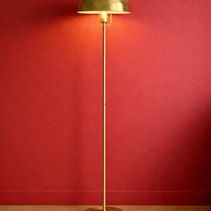 May include: A brass floor lamp with a domed shade, illuminating a warm glow against a red wall. The lamp has a slender pole and a circular base, creating a vintage aesthetic. The lamp is approximately 60 inches tall.