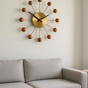 May include: A mid-century modern wall clock with a gold-colored face and brown wooden spheres marking the hours. The clock hands are black, with a red second hand. The clock is mounted on a white wall above a gray sofa.