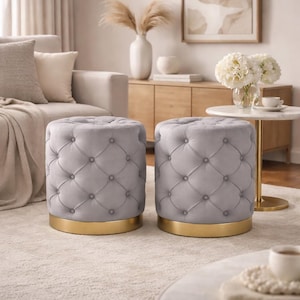 May include: Two grey, button-tufted ottomans with gold-coloured bases. The ottomans are cylindrical and placed on a light-coloured rug. The button detailing adds a touch of elegance. These ottomans are a stylish and functional piece of furniture for a living room.