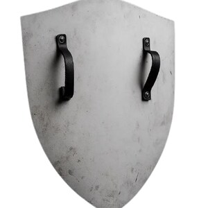 K&ouml;nnte beinhalten: Ein schildf&ouml;rmiges Objekt mit hellgrauer Oberfl&auml;che. Zwei schwarze Ledergriffe sind mit Nieten auf der R&uuml;ckseite befestigt. Das Schild hat ein leicht abgenutztes Aussehen, was auf Alter oder Gebrauch hindeutet.