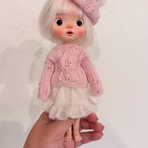 May include: A doll with short white hair wearing a pink knitted sweater with sequin accents, a matching pink beret with a white pom-pom, and a white ruffled skirt. The doll has large black eyes, rosy cheeks, and a small mouth on a pale complexion.