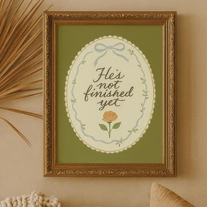 Puede incluir: Una impresión enmarcada con un marco dorado y ornamentado. La impresión tiene un fondo verde con un diseño ovalado en el centro. El óvalo contiene el texto "He's not finished yet" y una ilustración de rosa.
