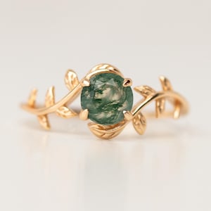 Könnte beinhalten: Ein goldfarbener Ring mit einem ovalen, grünen Stein in der Mitte. Der Ring hat ein zartes Blatt- und Rankenmuster, das den Stein umgibt. Der Ring ist vor einem weißen Hintergrund platziert.