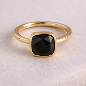 Könnte beinhalten: Ein goldfarbener Ring mit einem quadratischen, schwarzen Edelstein. Der Ring hat ein schlichtes Band und eine Lünettenfassung, die den dunklen Stein einfasst. Der Ring wird auf einer hellen, strukturierten Oberfläche präsentiert.