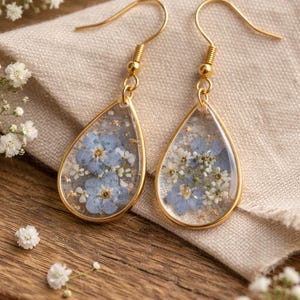 Könnte beinhalten: Ein Paar goldfarbene Tropfenohrringe. Jeder Ohrring hat einen Anhänger aus klarem Harz mit blauen und weißen Trockenblumen und Goldflocken. Die Ohrringe hängen an goldfarbenen Ohrhaken. Der Hintergrund ist Holz und weiße Blumen.