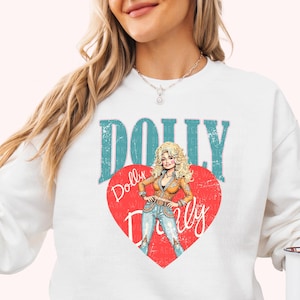 A woman wearing a white sweatshirt with a graphic design featuring a cartoon illustration of a blonde woman with curly hair and the word "Dolly" written on it.