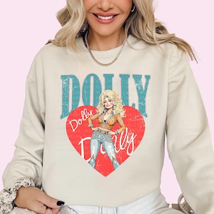 A woman wearing a white sweatshirt with a graphic design featuring a cartoon illustration of a blonde woman with curly hair and the text "Dolly Dolly" on it.