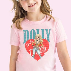 A young girl wearing a pink t-shirt with a graphic of a woman and the text "Dolly Dolly" printed on it.
