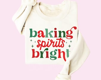 May include: Cream-colored sweatshirt with the text "baking spirits bright" in red and green. The sweatshirt is folded, with the sleeves and collar visible. The background is a light pink.