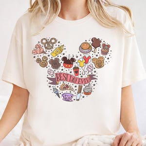 A white t-shirt with a Mickey Mouse-themed design featuring various Disney-related items such as ice cream cones, hot dogs, and other iconic characters.