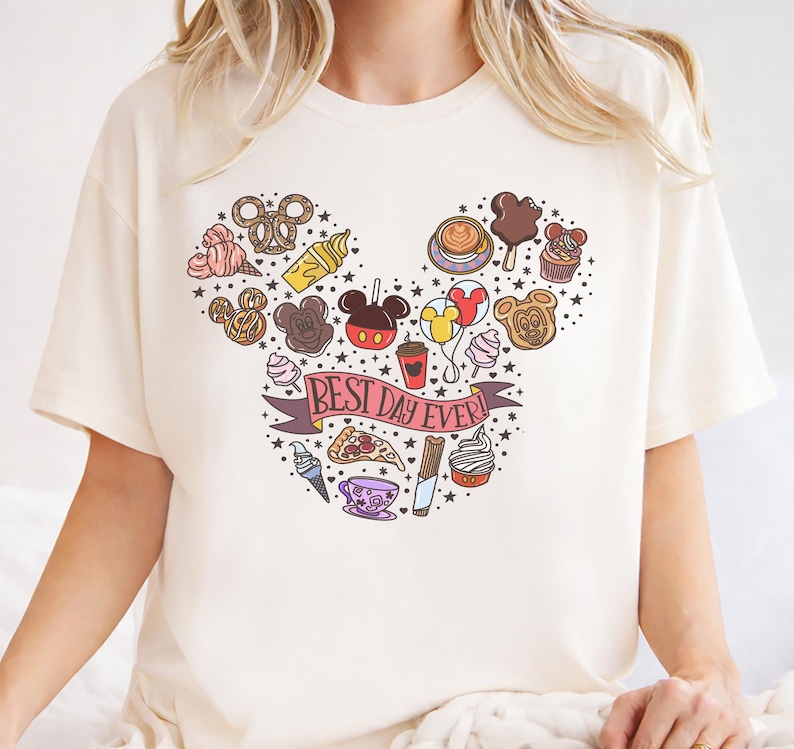 A white t-shirt with a Mickey Mouse-themed design featuring various Disney-related items such as ice cream cones, hot dogs, and other iconic characters.
