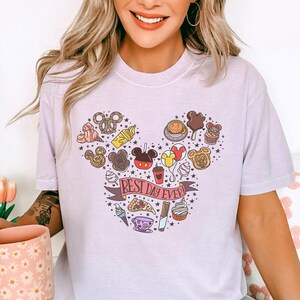 A woman wearing a white t-shirt with a graphic design featuring various Disney-themed items and the text "Best day ever!"