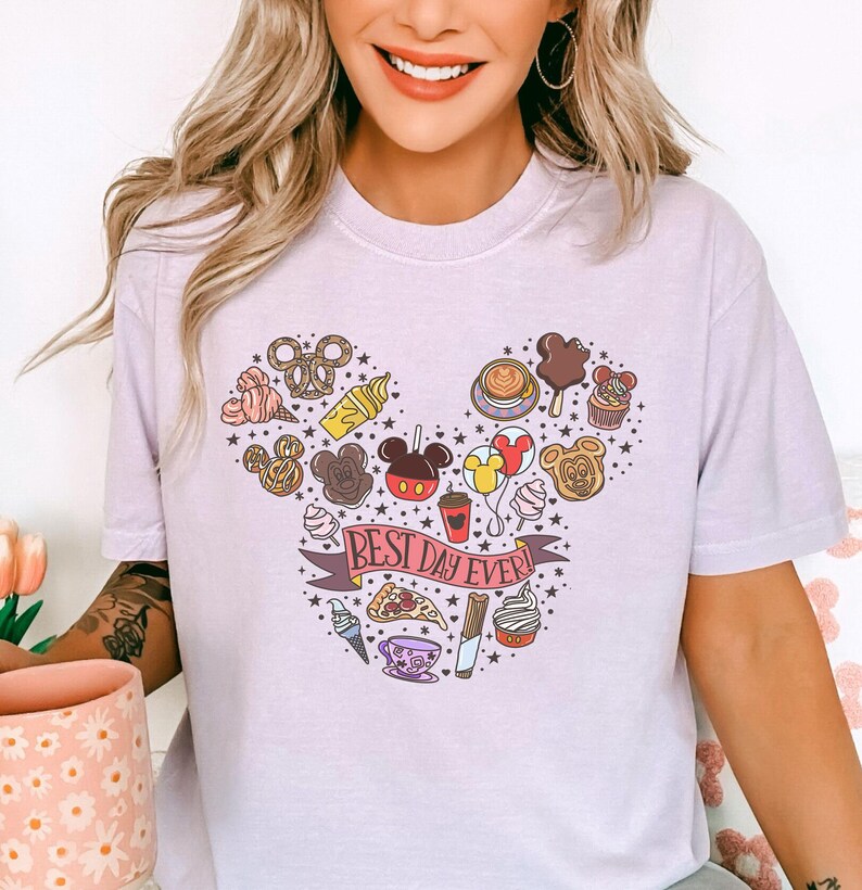 A woman wearing a white t-shirt with a graphic design featuring various Disney-themed items and the text "Best day ever!"
