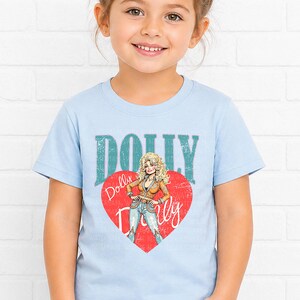 A young girl wearing a light blue t-shirt with a graphic of a woman and the text "Dolly Dolly" printed on it.