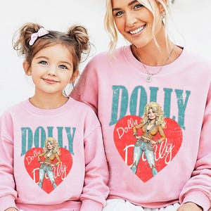 A young girl and an adult woman wearing matching pink sweatshirts with the word "Dolly" printed on them.