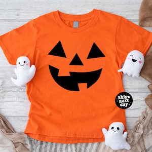 May include: Orange t-shirt with a black jack-o'-lantern face design. The shirt has a round neck and short sleeves. Three small white ghost plushies with smiling faces are placed around the shirt. The text "ships next day" is on a black circle.