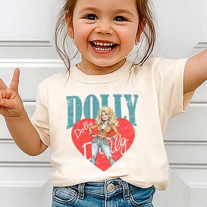 A young girl wearing a white t-shirt with a graphic of Dolly Dolly on it, smiling and making a peace sign.