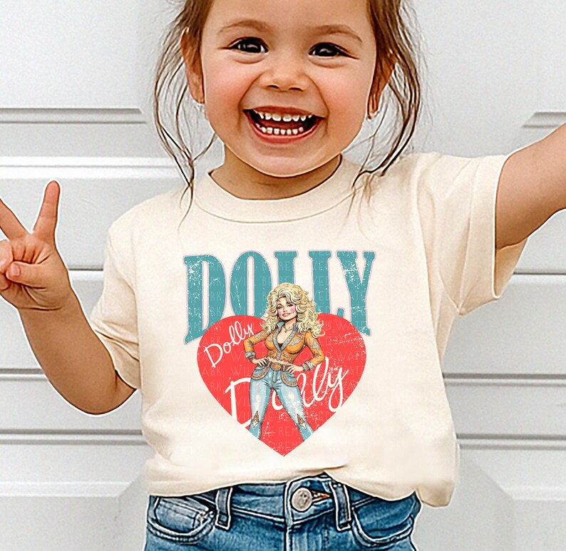 A young girl wearing a white t-shirt with a graphic of Dolly Dolly on it, smiling and making a peace sign.
