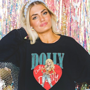 A woman wearing a black shirt with a graphic of a blonde woman and the word "Dolly" printed on it, posing in front of a colorful, glittery background.