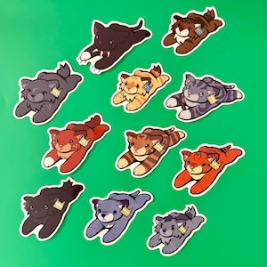 May include: A collection of ten illustrated stickers featuring various cats in a prone, pouncing position. Each cat has a distinct color pattern, including brown, gray, orange, and black, and each wears a small, rectangular tag on its ear.