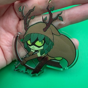 May include: A clear acrylic keychain featuring a cartoon character with a green and brown color scheme. The character has a hood, antlers, and is holding a bow and arrow. The keychain has a gold-colored clasp.