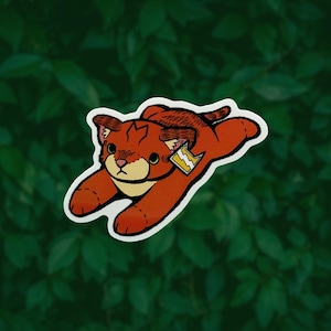 May include: A die-cut sticker featuring a stylized orange cat in a pouncing position. The cat has a cream-colored muzzle, dark brown ear markings, and a small tag attached to its ear displaying a yellow lightning bolt symbol on a white background.