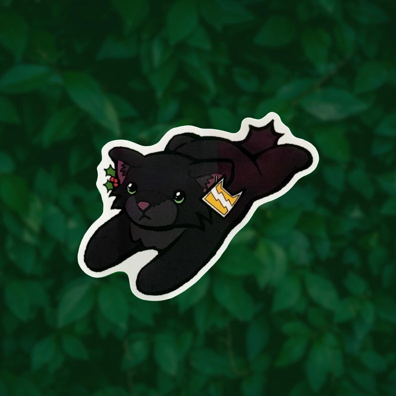 May include: A sticker featuring a black cat in a prone position with large green eyes and a pink nose. The cat has a small sprig of holly with red berries near its ear and a yellow tag with a white lightning bolt symbol attached to its side.