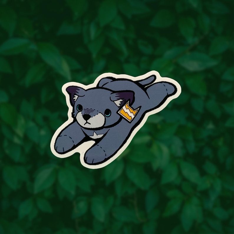 May include: A sticker featuring a cartoon-style gray cat in a pouncing position. The cat has dark gray ears, a light gray muzzle, and a white chest patch. A small orange tag with a white lightning bolt symbol is attached to the cat's ear.