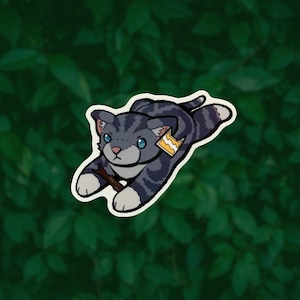 May include: A sticker illustration of a grey tabby cat with dark stripes, blue eyes, and a pink nose, shown in a pouncing position while holding a small brown twig. The cat wears a yellow tag with a white lightning bolt symbol on its collar.