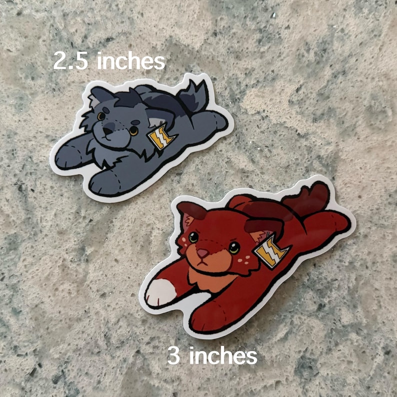 May include: Two die-cut stickers featuring stylized plush-like creatures in a prone position. The top sticker is dark gray and measures 2.5 inches, while the bottom sticker is deep red with white paw tips and measures 3 inches. Both have yellow tags.