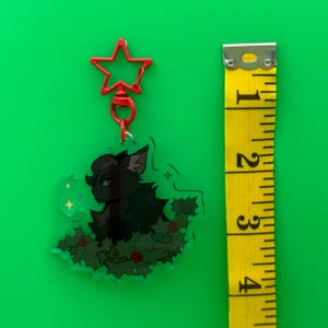 Puede incluir: Un charm transparente de acr&iacute;lico con un gato negro rodeado de hojas verdes de acebo y bayas rojas. Se adjunta a un cierre rojo en forma de estrella. El charm mide aproximadamente 5 cm de altura.