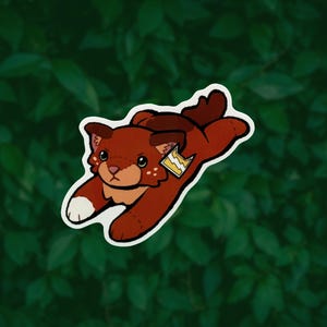 May include: A sticker featuring a brown, plush-style cat character in a pouncing pose. The cat has a white paw tip, light brown facial markings, and a small yellow tag attached to its ear with a white lightning bolt symbol printed on it.