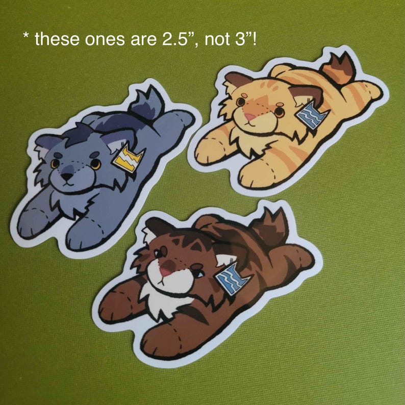 May include: Three die-cut stickers featuring illustrated plush-style cats in gray, orange, and brown. Each cat has a small tag on its ear. The text at the top reads: * these ones are 2.5", not 3"!. The stickers are arranged on a solid green surface.