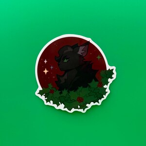 Pu&ograve; includere: Un adesivo raffigurante un gatto nero con occhi verdi, su uno sfondo rosso con stelle. Il gatto &egrave; circondato da agrifoglio verde con bacche rosse. L'adesivo ha un bordo bianco e si trova su uno sfondo verde.