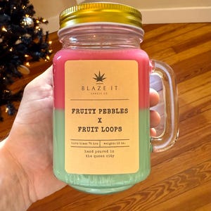 May include: A jar-shaped candle with a gold lid, featuring pink and green layers. The label reads "FRUITY PEBBLES x FRUIT LOOPS" and "BLAZE IT CANDLE CO." The candle weighs 15 oz and has a burn time of 76 hours.